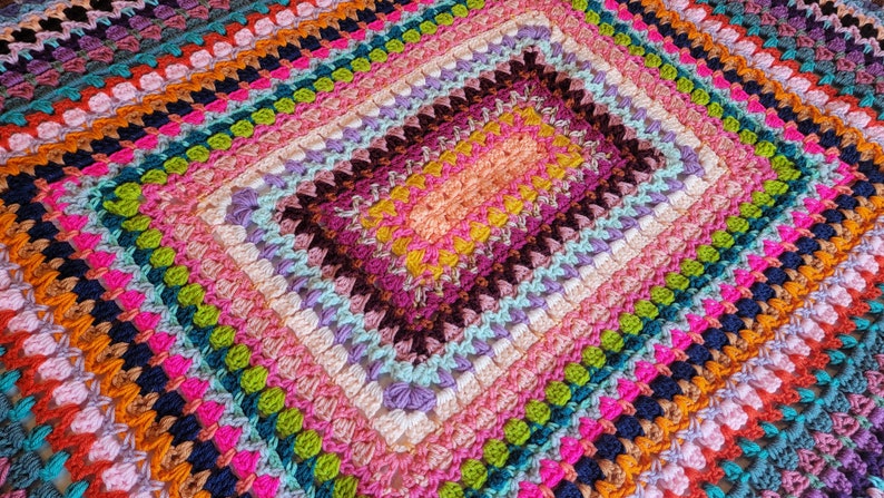 May include: A colourful crocheted blanket with a square pattern in the centre. The blanket is made with a variety of colours, including pink, purple, green, blue, and orange.