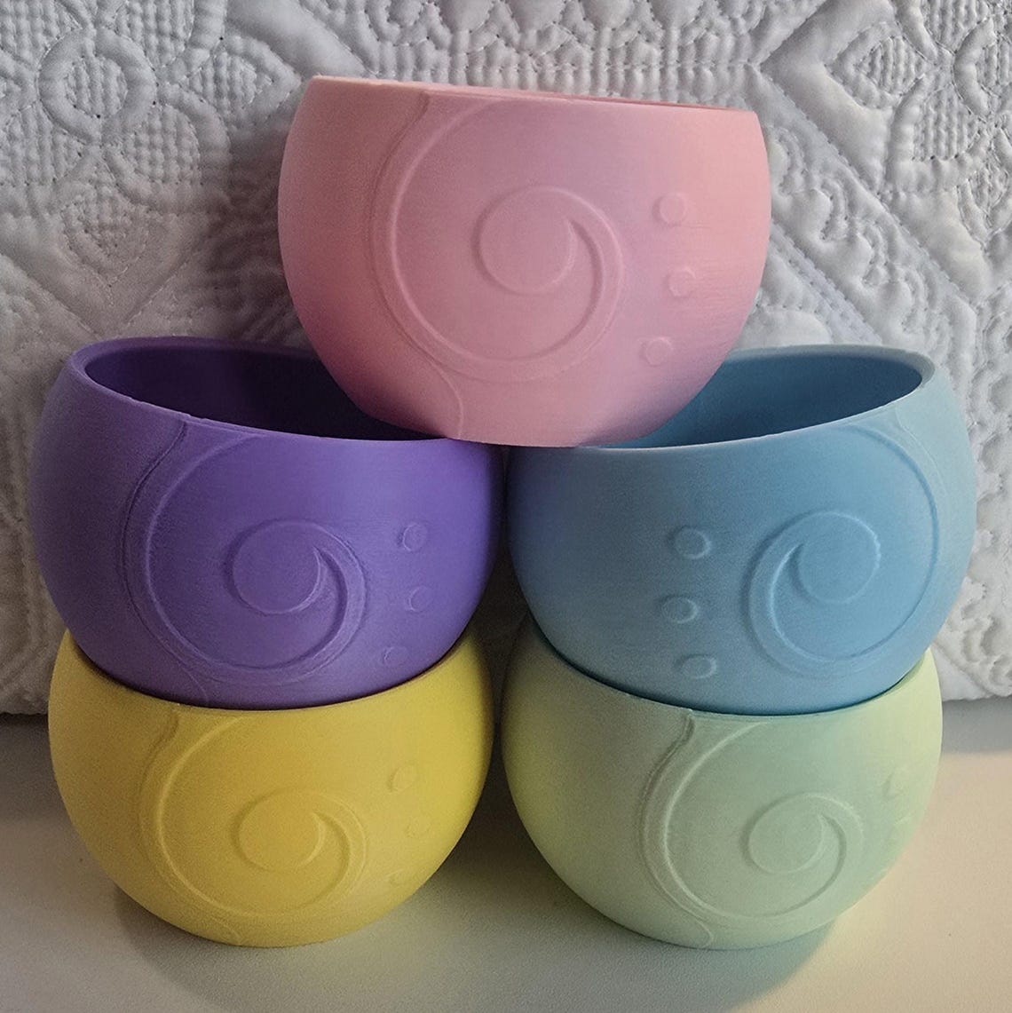 May include: Four silicone nesting bowls in pastel colors. The bowls are stacked on top of each other. The top bowl is pink, the second bowl is purple, the third bowl is yellow, and the bottom bowl is green. Each bowl has a swirl design and small dots around the swirl.