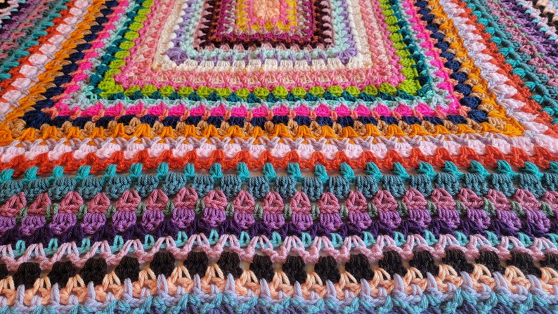 May include: A colourful crocheted blanket featuring a range of stitches and colours, including pink, orange, green, blue and purple.