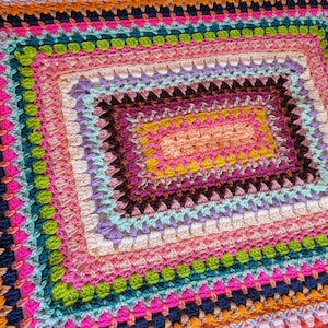 May include: A colourful crocheted blanket with a multi-coloured square pattern in the centre. The blanket is made with a variety of different coloured yarns, including pink, purple, blue, green, yellow, orange, and red.