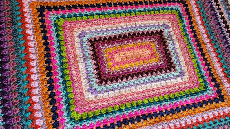 May include: A colourful crocheted blanket with a multi-coloured square pattern in the centre. The blanket is made with a variety of different coloured yarns, including pink, purple, blue, green, yellow, orange, and red.
