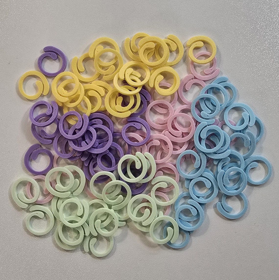 May include: A large pile of colorful plastic stitch markers in yellow, light green, light blue, and purple. The markers are in the shape of a circle with a small opening in the center.