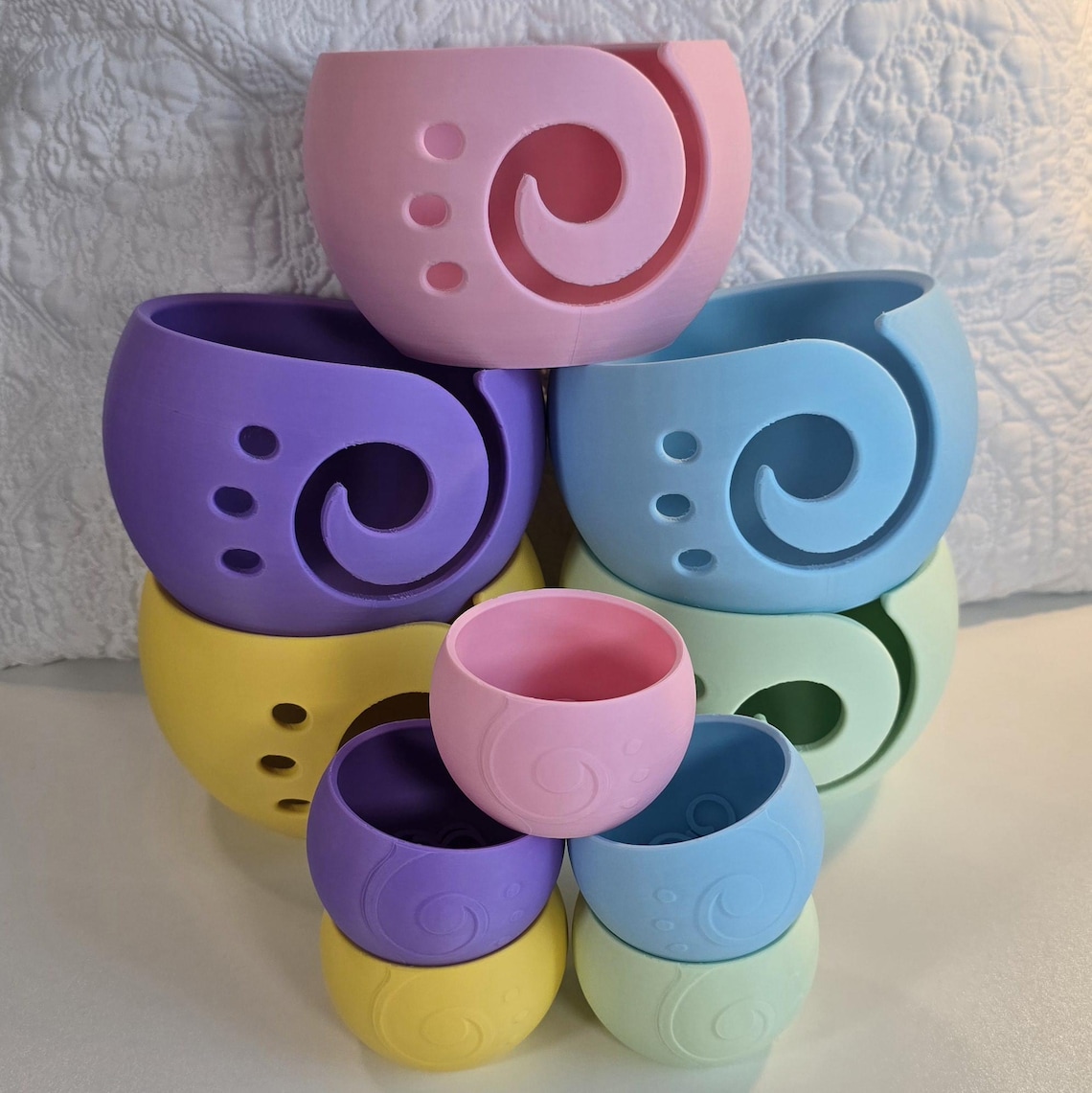 May include: A stack of six colorful silicone yarn bowls. The bowls are in various shades of purple, blue, yellow, and pink. Each bowl has a unique swirl design and holes for yarn to pass through.