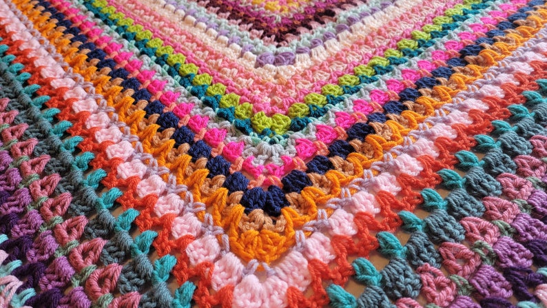 May include: A colourful crocheted blanket featuring a range of bright colours, including pink, orange, green, blue, and purple. The blanket is crafted with a textured stitch that creates a unique and interesting pattern.