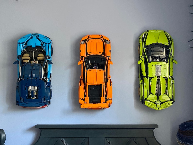 Może przedstawiać: Trzy modele samochod&oacute;w Lego wystawione na ścianie. Samochody to niebieski Bugatti Chiron, pomarańczowe Porsche 911 GT3 RS i zielony Lamborghini Aventador SVJ.