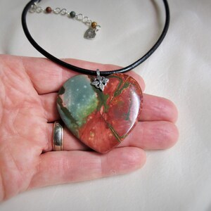 May include: A heart-shaped pendant necklace with a green, red, and brown stone. The pendant is attached to a black leather cord.