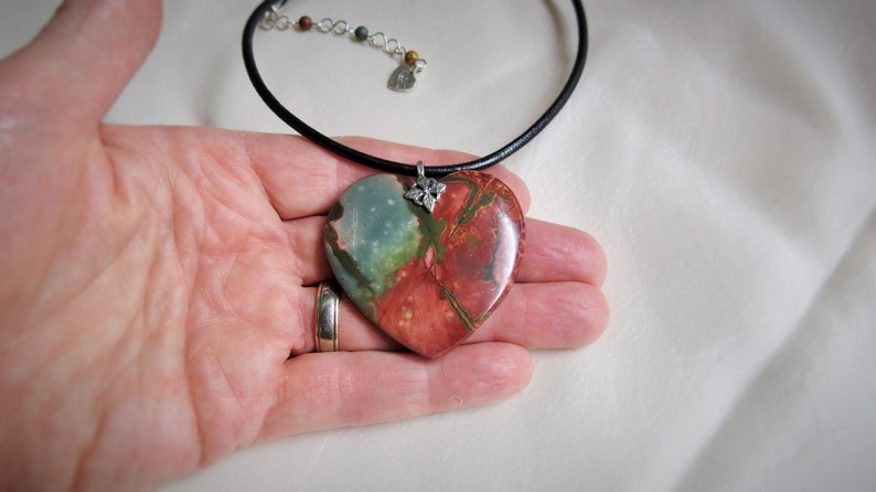 May include: A heart-shaped pendant necklace with a green, red, and brown stone. The pendant is attached to a black leather cord.