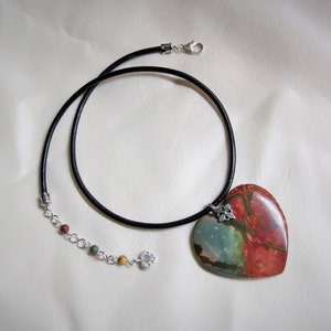 May include: A heart-shaped pendant necklace with a red, green, and blue stone. The necklace has a black leather cord and a silver chain with small beads.