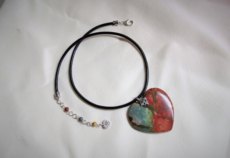 May include: A heart-shaped pendant necklace with a red, green, and blue stone. The necklace has a black leather cord and a silver chain with small beads.