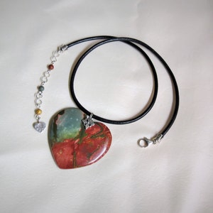 May include: A heart-shaped pendant necklace with a red, green, and blue stone on a black leather cord. The pendant is attached to a silver chain with small silver beads.