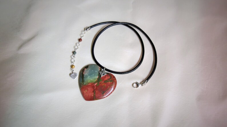 May include: A heart-shaped pendant necklace with a red, green, and blue stone on a black leather cord. The pendant is attached to a silver chain with small silver beads.