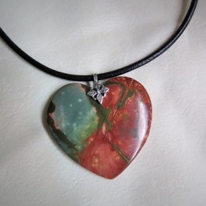 May include: A heart-shaped pendant necklace with a black leather cord. The pendant is made of a green, red, and brown stone with a silver flower charm.