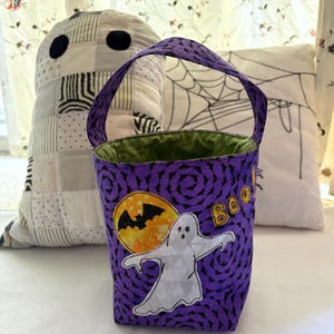 May include: A purple fabric treat bag featuring a repeating bat pattern, a white ghost applique, a yellow moon with a black bat silhouette, and the word BOO in yellow letters. The bag has a green interior lining and a single purple fabric handle.