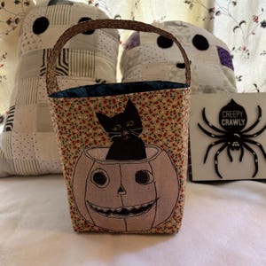 May include: A fabric basket with a handle featuring a small floral print in red, green, and yellow. The front displays a black cat sitting inside a light-colored pumpkin illustration with a carved face. The interior lining is a solid dark blue fabric.