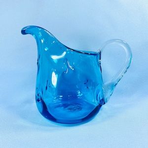 May include: A small, turquoise-blue glass creamer with a curved spout and a clear, looped handle. The creamer has a textured surface and sits on a light blue background. The design is reminiscent of vintage glassware.