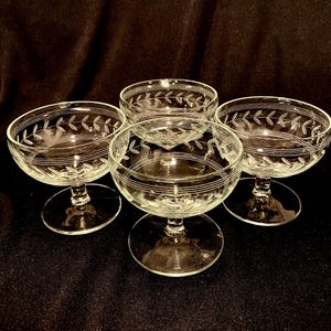 May include: Four clear glass champagne coupe glasses. Each glass features a delicate etched leaf and line design around the rim and bowl. The glasses have a stemmed base and are arranged on a dark surface.