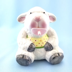May include: A ceramic figurine of a white lamb with a pink nose and ears, wearing a yellow bib with red polka dots. The lamb has a pink pacifier in its mouth and dark gray hooves on its feet, sitting in a forward-facing position.