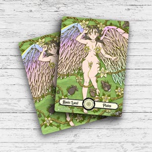 May include: Two trading cards featuring a nude woman with large feathered wings, surrounded by flowers and rabbits. The cards have a green background with the text "Basic Land" and "Plains". The woman has long dark hair and is adorned with flowers.
