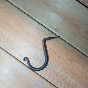 May include: A black metal hook with a simple design. The hook is curved and has a pointed end. It is shown against a wooden background.