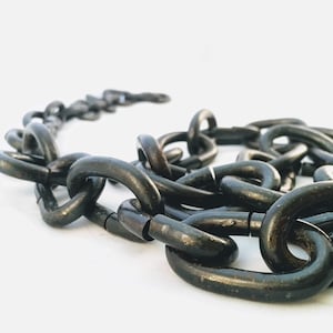 Puede incluir: Primer plano de una cadena de metal negro. La cadena est&aacute; hecha de eslabones gruesos e interconectados. La cadena est&aacute; sobre una superficie blanca.