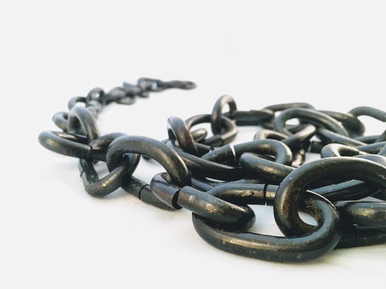 Puede incluir: Primer plano de una cadena de metal negro. La cadena est&aacute; hecha de eslabones gruesos e interconectados. La cadena est&aacute; sobre una superficie blanca.