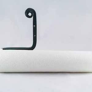 May include: Black metal paper towel holder with a decorative scroll design. The holder is mounted to a wall and has a white paper towel roll on it.