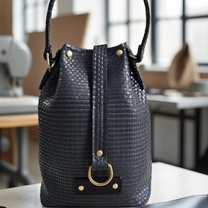 Può includere: Borsa a secchiello nera con trama intrecciata, manico intrecciato e dettagli dorati. La borsa ha una chiusura con coulisse e un dettaglio decorativo con anello a D. La borsa è appoggiata su un tavolo.