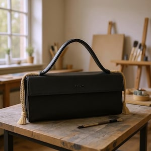 Puede incluir: Un bolso rectangular negro con un asa curva y detalles de cuerda. El bolso está sobre una mesa de madera con un pincel cerca. El bolso tiene un cierre de solapa y un pequeño logotipo.