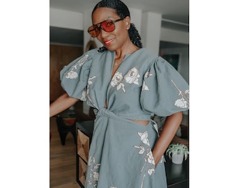 May include: A woman wearing a blue wrap top dress with a floral print. The dress has a tie at the waist and a cutout detail at the side. The dress is styled with a pair of red sunglasses. The text on the image reads "Donna Wrap Top Dress, PDF Sewing Pattern, Print at home, Style #8600, Size Small-XL".