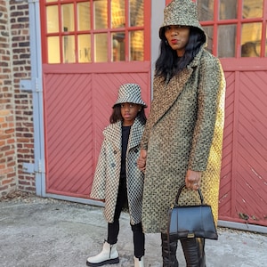 Puede incluir: Dos personas con abrigos y sombreros a juego con estampado negro y dorado. La persona de delante lleva botas blancas. La persona de atrás lleva un bolso de mano negro.