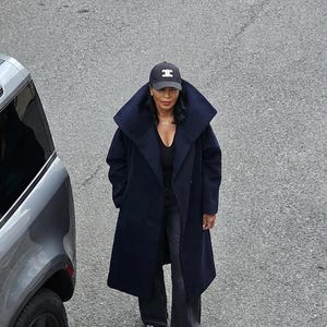 May include: A person wearing a navy blue coat and a black baseball cap with a white letter "H". The long coat has a wide collar. They are standing on a grey surface.