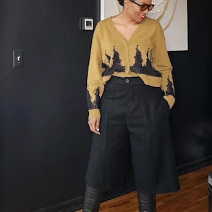 Könnte beinhalten: Eine Frau trägt einen senfgelben Cardigan mit schwarzen Spitzendetails, schwarze Culottes und schwarze kniehohe Stiefel.