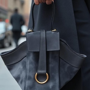 May include: A dark blue suede handbag with a gold ring detail and a flap closure. The bag has a structured shape and a top handle.
