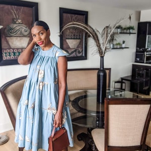 Puede incluir: Vestido azul claro escalonado con estampado floral. El vestido tiene mangas cortas y se combina con tacones marrones y un bolso marrón. El vestido llega a la rodilla y parece estar hecho de una tela ligera.
