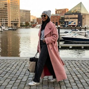 Puede incluir: Una persona con un abrigo largo rosa claro, gorro gris, gafas de sol y vaqueros negros camina por un camino adoquinado. Lleva un bolso tote negro con un panel beige. El fondo presenta una zona marítima con barcos y edificios.