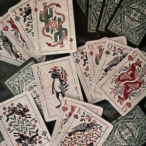 May include: A deck of playing cards with a botanical and animal theme. The cards feature illustrations of snakes, rabbits, birds, and flowers in red, green, and black on a cream background. The card backs are dark green with a floral design.