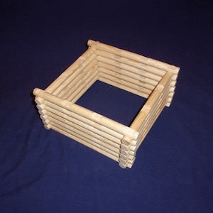 May include: A wooden log cabin model made of stacked wooden dowels. The model is unfinished and has a square shape.