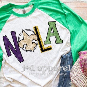 May include: A white and green raglan t-shirt with the word "NOLA" in colorful letters and a fleur-de-lis design. The shirt is on a wooden surface with a pair of jeans and a bedazzled hat.