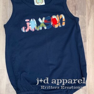 May include: A navy blue baby onesie with a colorful patchwork name "Jaxson" on the front. The onesie has a snap closure at the crotch and is made of soft, comfortable fabric.