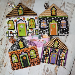 May include: Four Halloween-themed house-shaped pot holders. Each features a unique design with a door, windows, and a roof. The color palette includes orange, black, green, and purple, with spooky accents like ghosts and bats. Each has a loop for hanging.