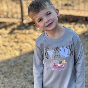 May include: A gray long-sleeved shirt with the name "JAXSON" in black and white letters with a clock design. The shirt also has a pink and white logo that says "Knitting Kreations".
