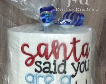 May include: A white toilet paper roll with red, black, and blue embroidery that reads "Santa said you are a cheap". The roll is wrapped in clear plastic with a blue ribbon.