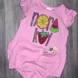 May include: Pink baby onesie with a ruffle trim and embroidered fruit design. The onesie says "Nora Kay" with a lemon, watermelon, and strawberry.