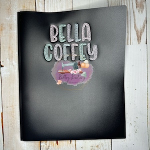 May include: A black notebook with the text "BELLA COFFEY" in a sparkly, multi-colored font. Below the text is a logo with a sewing machine, flowers, and the words "Krittys Kreations". The notebook is on a light wood surface.