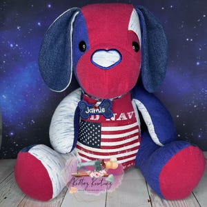 May include: A red, white, and blue patriotic dog plush toy with a name tag that says "Jannie" and a US Navy shirt. The dog is sitting on a dark blue background with white stars.