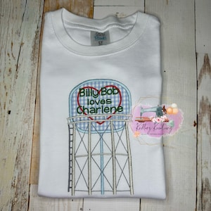 May include: White t-shirt with an embroidered design of a water tower with a heart-shaped sign that reads "Billy Bob loves Charlene".