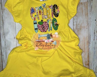 May include: Yellow dress with a colorful applique design that says "Emilie One" with school supplies and a sewing machine. The dress has a ruffled hem.