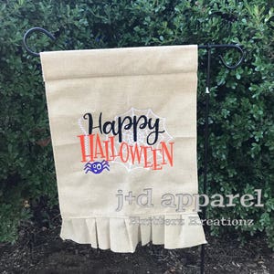 Könnte beinhalten: Eine Gartenflagge aus Burlap mit dem Text "Happy Halloween" in schwarzer und orangefarbener Schrift mit einem weißen Spinnennetz und einer Cartoon-Spinne. Die Flagge hat einen gekräuselten unteren Rand und der Text "j+d apparel Kritterz Kreations" ist unten in Grau gedruckt.