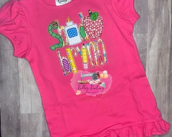 May include: Pink short-sleeved shirt with a ruffled hem. The shirt has a colorful applique design that says "School Brynn" with a worm, pencil, ruler, glue, and apple.