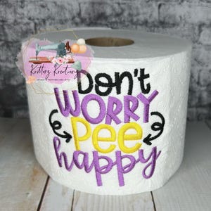 May include: A white toilet paper roll with purple, yellow, and black embroidery that reads "Don't Worry, Pee Happy".